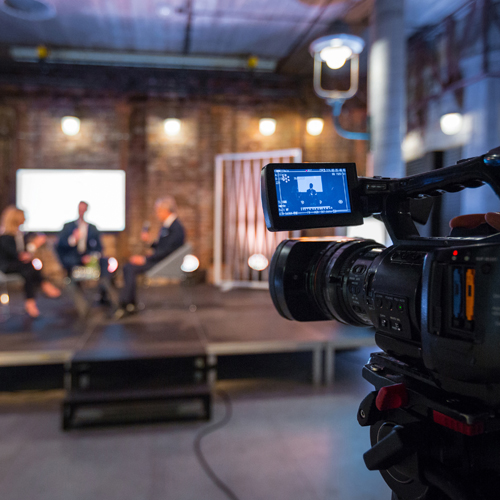 Businesswoman and businessman giving presentation during online seminar, sitting on armchairs on the stage, holding microphones. Focus on video camera on the foreground.